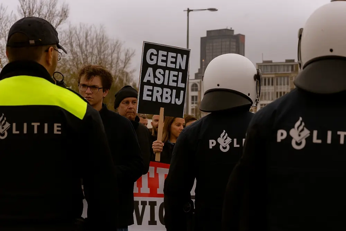 Drie Nederlandse politieagenten staan tegenover demonstranten in Amsterdam, waaronder een jongeman met bril en een protestbord met de tekst “GEEN ASIEL ERBIJ”, tegen een achtergrond van grijze lucht en stedelijke hoogbouw.